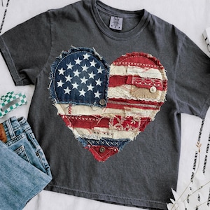 Könnte beinhalten: Ein anthrazitfarbenes T-Shirt mit einem herzförmigen Design. Das Design besteht aus Flicken aus Denim, rotem und weißem Stoff und Jute. Die linke Seite des Herzens zeigt einen blauen Denim-Flicken mit weißen Sternen, die rechte Seite rote und weiße Streifen.
