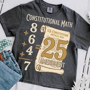 May include: Dark gray t-shirt with the words "Constitutional Math" and a math problem with the answer 25. A scroll graphic reads "US Constitution Article XXV 25 Amendment". The shirt is paired with blue jeans.