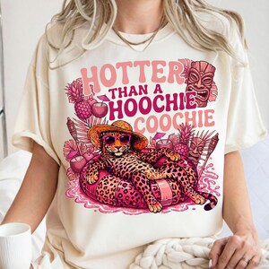 May include: Cream-colored t-shirt with a graphic design featuring a leopard wearing sunglasses and a straw hat, lounging on a pink inflatable. The text reads "Hotter Than a Hoochie Coochie" in pink. Tropical elements like pineapples and tiki masks surround the leopard.