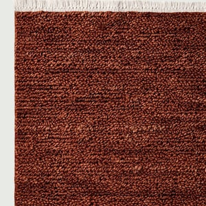 Puede incluir: Primer plano de una alfombra texturizada de color óxido con un borde de flecos blancos. La superficie de la alfombra está densamente tejida, mostrando un tono rico y terroso. Esta alfombra es adecuada para la decoración del hogar.