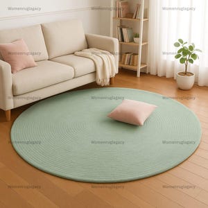 Könnte beinhalten: Ein runder, hellgrüner Teppich mit konzentrischem Design. Ein hellrosa quadratisches Kissen liegt auf dem Teppich. Ein beiges Sofa und eine kleine Topfpflanze befinden sich im Hintergrund. Der Teppich liegt auf einem Holzboden.