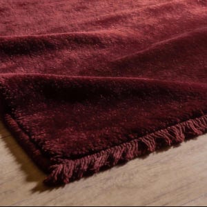 May include: Close-up of a deep burgundy rug with a fringed edge. The rug is folded over, showing its thick texture and rich colour. The rug is on a light-coloured wooden floor.