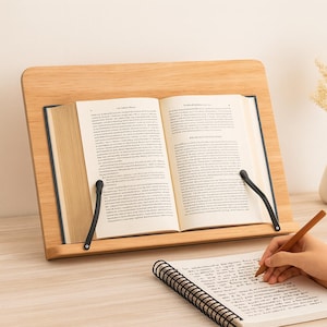 May include: A wooden book stand with an open book and black page holders. A spiral notebook and a person's hand holding a pen are in the foreground. The book stand is made of light-colored wood.