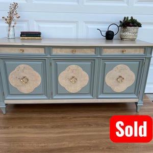 May include: A light blue and beige wooden cabinet with three doors and drawers. The cabinet has gold hardware and decorative floral accents. A small black watering can and potted plant sit on top. The word "Sold" is in a red box.