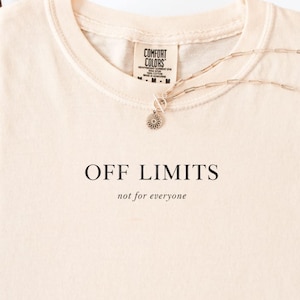 May include: Ivory-colored t-shirt with the text "OFF LIMITS" and "not for everyone" in black. A gold-colored necklace with a pendant rests on the shirt. The shirt is folded neatly on a white surface.
