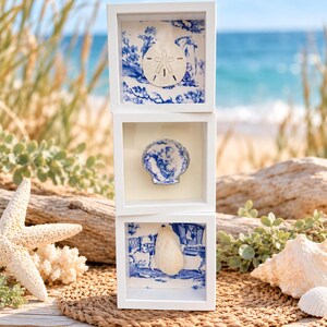 May include: A vertical stack of three white square shadow box frames. Each frame contains a white seashell mounted against a blue and white toile patterned background featuring pastoral scenes of figures and carriages in a classic style.