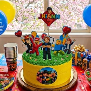 以下が含まれることがあります： 「Jhiann 7」と書かれたトッパーと、Robloxのキャラクターフィギュアで飾られた黄色の誕生日ケーキ。ケーキは木製のケーキスタンドに置かれ、パーティー用品、風船、プレゼントに囲まれています。