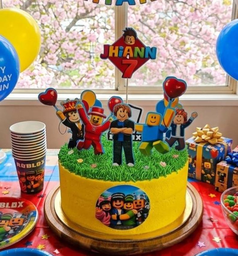 以下が含まれることがあります： 「Jhiann 7」と書かれたトッパーと、Robloxのキャラクターフィギュアで飾られた黄色の誕生日ケーキ。ケーキは木製のケーキスタンドに置かれ、パーティー用品、風船、プレゼントに囲まれています。