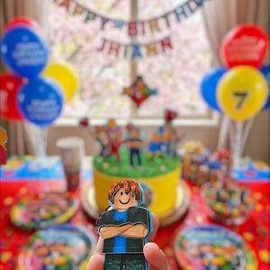 以下が含まれることがあります： ロブロックスをテーマにしたケーキ、風船、装飾が施された誕生日パーティーのシーン。ロブロックスのキャラクターの切り抜きが前景に保持されています。「Happy Birthday Jhiann」と書かれたバナー。テーブルにはお皿とお菓子が並んでいます。
