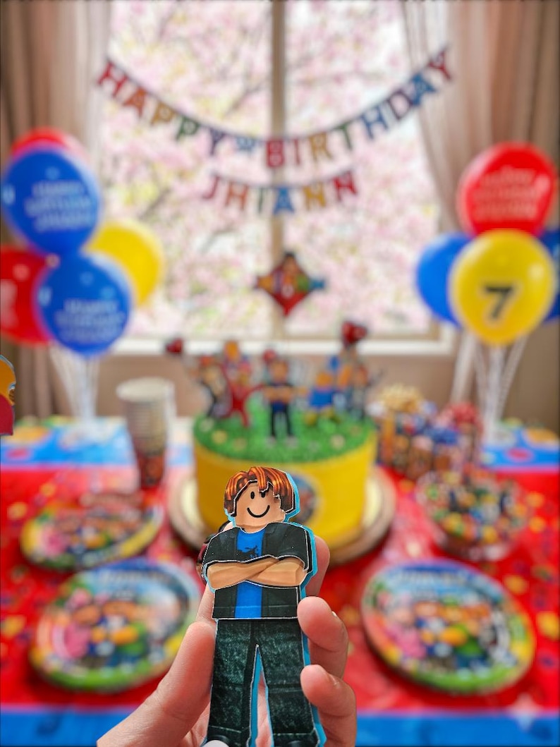 以下が含まれることがあります： ロブロックスをテーマにしたケーキ、風船、装飾が施された誕生日パーティーのシーン。ロブロックスのキャラクターの切り抜きが前景に保持されています。「Happy Birthday Jhiann」と書かれたバナー。テーブルにはお皿とお菓子が並んでいます。