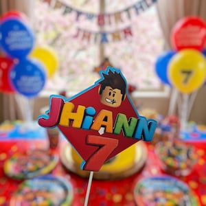 以下が含まれることがあります： 漫画のキャラクターと「Jhiann」の名前と数字「7」が付いたバースデーケーキトッパー。トッパーは赤、青、黄、オレンジ色です。「Happy Birthday Jhiann」と書かれた風船とバナーが背景にあります。