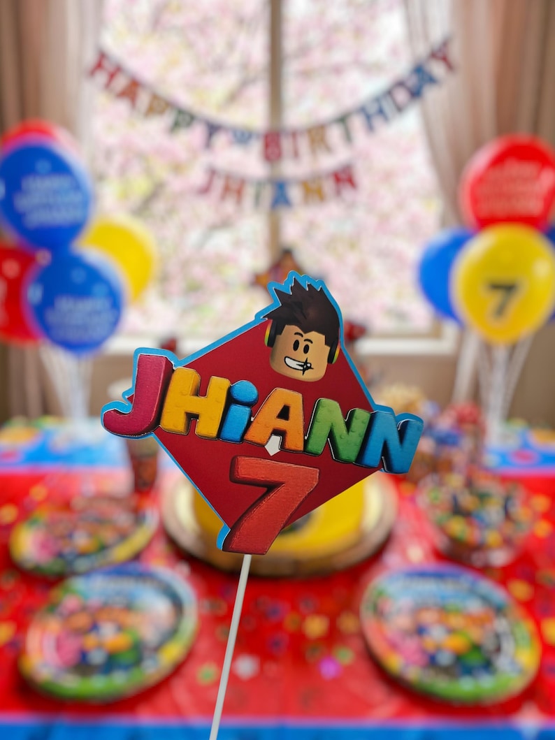 以下が含まれることがあります： 漫画のキャラクターと「Jhiann」の名前と数字「7」が付いたバースデーケーキトッパー。トッパーは赤、青、黄、オレンジ色です。「Happy Birthday Jhiann」と書かれた風船とバナーが背景にあります。