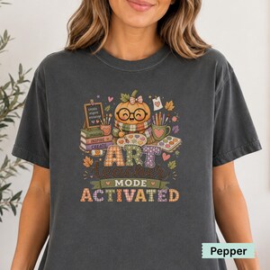 May include: A charcoal gray t-shirt featuring a graphic of a pumpkin wearing glasses and a scarf, painting with a brush. The design includes books labeled "IMAGINE," "INSPIRE," "CREATE," a chalkboard with "create inspire encourage," and the text "ART TEACHER MODE ACTIVATED."