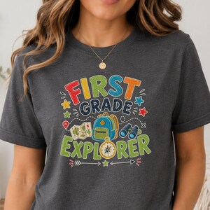 May include: A dark charcoal gray t-shirt featuring a colorful graphic with the text FIRST GRADE EXPLORER. The design includes a blue backpack, a map, binoculars, and a compass, surrounded by small stars and dashed lines in red, yellow, and green.