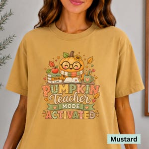 May include: A mustard yellow t-shirt featuring a graphic of a smiling pumpkin wearing glasses and a scarf, surrounded by books labeled Teach, Inspire, Love, a mug, and autumn leaves. The text reads PUMPKIN Teacher MODE ACTIVATED.