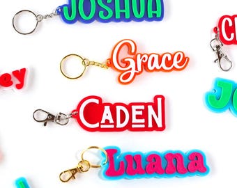 May include: A collection of colorful name keychains in various colors, including blue, green, red, and orange. Each keychain features a different name in a bold, playful font. Some have key rings, others have lobster clasps.