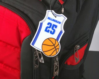 Puede incluir: Mochila negra y roja con una etiqueta de camiseta de baloncesto. La etiqueta es blanca con ribete azul y presenta el nombre "Dillingham" y el número "25", junto con un gráfico de baloncesto. La mochila tiene múltiples compartimentos con cremallera.