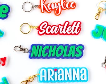 May include: Keychains with names in various colors, including red, orange, blue, and green. Each keychain has a metal ring or clasp. Names include Kaylee, Scarlett, Nicholas, Arianna, and Grace.