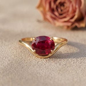 Puede incluir: Un anillo de oro con una gran piedra preciosa de color rojo intenso y corte ovalado, sujeta por cuatro garras. La banda tiene un diseño liso y curvado que se estrecha hacia el engaste central, mostrando un acabado metálico pulido y brillante.
