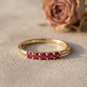 May include: A gold ring featuring a row of five small, round, vibrant red rubies set in a prong setting. The band is smooth and polished, showcasing a classic design that highlights the deep crimson color of the gemstones in a horizontal line.
