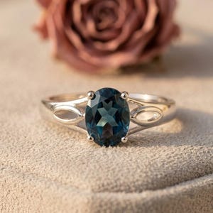 Puede incluir: Un anillo de plata con una piedra preciosa azul oscuro de corte ovalado sujeta por cuatro garras. La banda tiene un diseño dividido con aberturas en forma de ojo a cada lado de la piedra, creando un aspecto simétrico con acabado pulido.