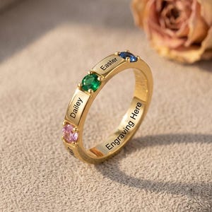 Puede incluir: Un anillo de tono dorado con tres piedras preciosas de corte redondo en rosa, verde y azul. La banda está grabada con los nombres Dailey y Easter, junto con el texto Engraving Here, situados entre las piedras y a lo largo de la banda interior.