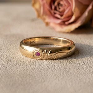 May include: A gold band ring featuring a small, round, pink gemstone set into the metal. The band is engraved with a cursive letter M and has the text 14K stamped on the interior surface. The ring has a polished, smooth, metallic finish.