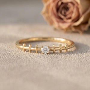 May include: A gold ring featuring a thin band with a series of vertical bar accents along the shank and a single round brilliant-cut white gemstone set in the center. The metal has a polished finish and a warm yellow tone throughout the design.
