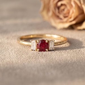 May include: A gold ring featuring a central round-cut red ruby flanked by two clear baguette-cut diamonds. The stones are set in a polished gold band, creating a three-stone design that highlights the contrast between the red and white gems.