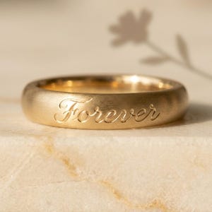May include: A gold band ring with a brushed matte finish featuring the word Forever engraved in a cursive script font on the outer surface. The ring has a rounded profile and a warm yellow gold tone, showcasing a clean and simple design.