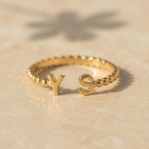 Puede incluir: Un anillo abierto de tono dorado con un diseño de banda de cuentas. Los dos extremos del anillo terminan con las letras mayúsculas Y y S, posicionadas frente a frente sobre la abertura de la banda, creando una pieza de joyería con iniciales.