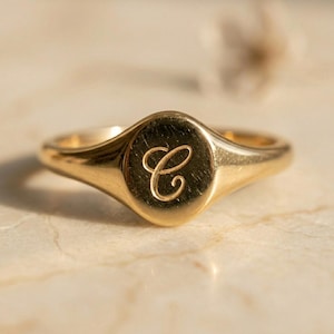 May include: A gold signet ring featuring a polished, smooth surface with a single, engraved cursive letter C centered on the oval face. The band is solid gold with a rounded profile, showcasing a reflective finish and clean, simple lines.