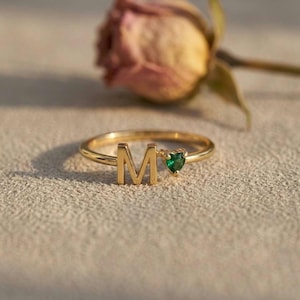 May include: A gold-toned ring featuring the capital letter M and a small, heart-shaped green gemstone. The band is thin and circular, showcasing a polished metallic finish that reflects light, positioned near a dried, muted pink rose.