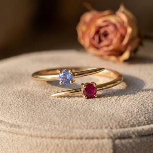 May include: A gold bypass ring featuring two round-cut gemstones, one light blue and one deep red, set in a prong setting. The ring has a slender, polished gold band that overlaps slightly at the centre, showcasing the contrasting coloured stones.