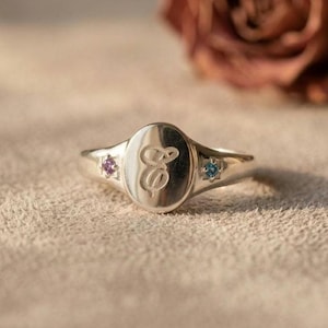 May include: A silver-toned signet ring featuring an oval face engraved with a cursive letter E. The ring band is accented on each side by a small, round, prong-set gemstone, with a purple stone on the left and a blue stone on the right side.