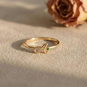 Puede incluir: Un anillo de tono dorado que presenta una letra A en cursiva como punto focal central. La letra A está acentuada con una pequeña piedra preciosa redonda de color verde vibrante engastada en el metal. El aro es fino, liso y circular.