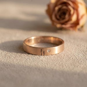 Peut inclure: Une bague anneau en or rose avec une finition brossée, gravée de deux petites silhouettes de pins et d'une unique pierre précieuse blanche ronde sertie à fleur, positionnée à droite des arbres sur la surface extérieure de l'anneau.