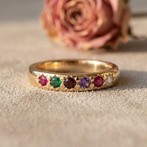 May include: A gold band ring featuring six small, round, multi-colored gemstones set in a row. The stones include a pink, green, dark red, purple, red, and clear gem, each embedded within a star-shaped metal setting along the outer surface.