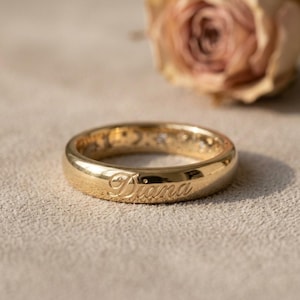 May include: A polished gold band ring featuring the name Diana engraved in a cursive script on the outer surface. The ring has a smooth, rounded profile and a reflective metallic finish, showcasing a clear and legible inscription on the front.
