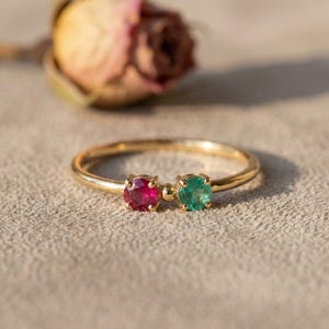 May include: A thin gold ring featuring two round-cut gemstones, one deep red and one vibrant green, set side-by-side with a small gold bead between them. The band is smooth and polished, showcasing a delicate design with prong settings.