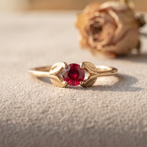 May include: A gold ring featuring a round, deep red gemstone set in the centre. The band is designed with two symmetrical, leaf-shaped metal prongs that curve inward to hold the stone securely in place, creating a botanical-inspired aesthetic.