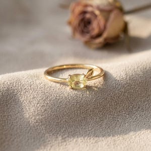 May include: A gold ring featuring a single oval-shaped pale yellow gemstone set in a prong setting. The band includes a small, smooth gold leaf-shaped accent attached directly next to the gemstone setting, creating a delicate and refined appearance.
