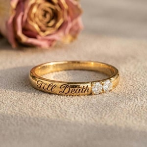 May include: A gold band ring featuring the engraved text Till Death in a cursive script. The ring is accented with two small, round, clear, faceted stones set side by side next to the lettering, creating a polished and reflective appearance.