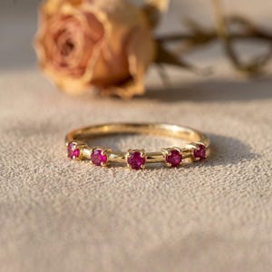 May include: A gold band ring featuring five small, round, vibrant pinkish-red gemstones set in individual prongs along the top half of the band. The ring has a polished metallic finish and a simple, slender design with a consistent circular shape.