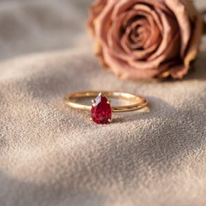 May include: A gold ring featuring a single pear-shaped red gemstone set in a prong setting. The band is thin and polished, showcasing a vibrant crimson stone that tapers to a point, reflecting light across its faceted surface.