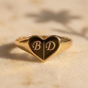 Puede incluir: Un anillo de sello en forma de corazón hecho de oro con un acabado liso y pulido. El centro del corazón está grabado con las letras mayúsculas B y D, separadas por una línea vertical. El anillo tiene una banda sólida y superficie metálica.