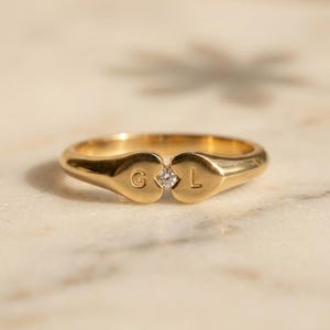 Puede incluir: Un anillo de oro con dos extremos en forma de corazón que se unen en el centro. El corazón izquierdo tiene grabada la letra G y el derecho la letra L. Una pequeña piedra preciosa redonda y transparente está engastada entre ambos.