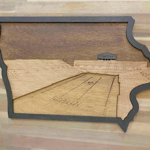 May include: Wooden wall art shaped like the state of Iowa, featuring a detailed stadium scene. The artwork has a dark gray border and a natural wood finish, with the word "IOWA" visible on the field.