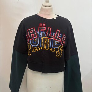 Può includere: Felpa corta nera con maniche lunghe verdi. La parte anteriore della felpa presenta la scritta "RÄELLE PARIS" in un design ricamato colorato. La felpa è esposta su un manichino.