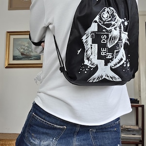 Può includere: Zaino nero con coulisse con grafica di pesce bianco. Il design del pesce include il testo "RE DS" e altri elementi testuali. Lo zaino è indossato sopra una camicia bianca e jeans blu.